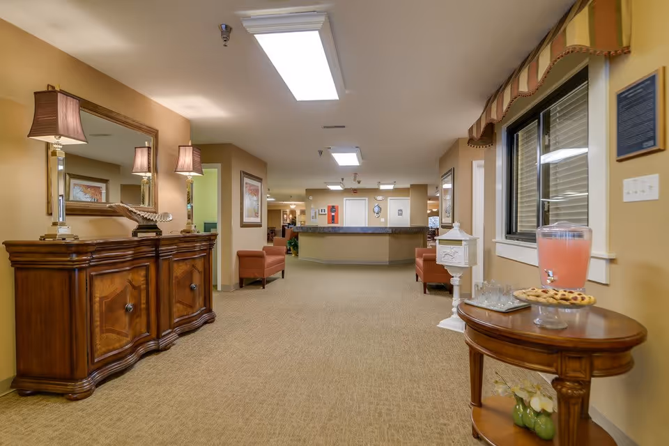 Interior view of a senior living facility hallway with beige walls and carpet. On the left, there is a wooden sideboard with two lamps and a large mirror above it. Two upholstered chairs are placed along the walls. On the right, a small wooden table holds a pitcher of pink lemonade, glasses, and a plate of cookies. A window with blinds is above the table. In the background, a reception desk is visible with doors and framed pictures on the walls.