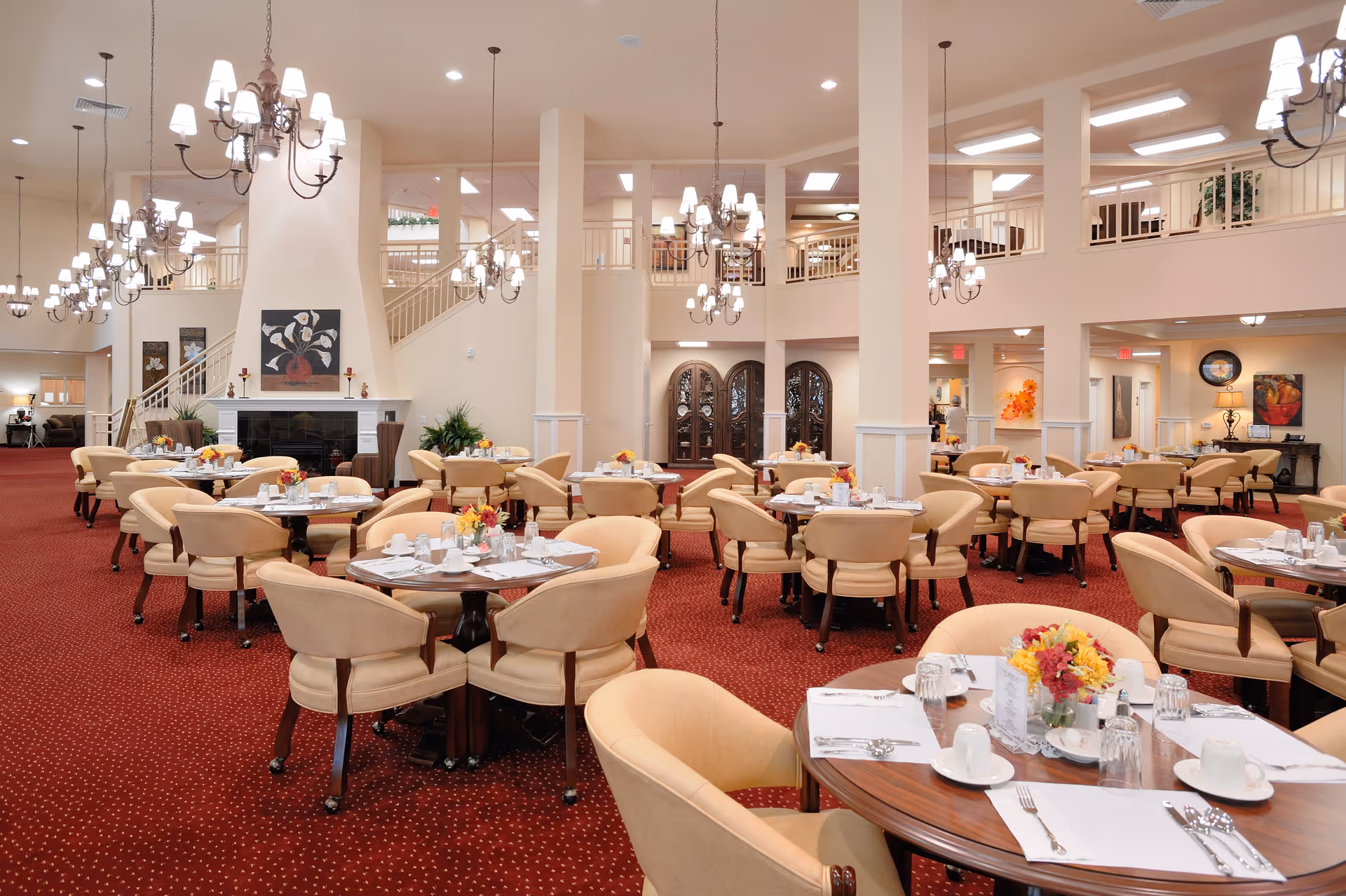 A spacious and elegant dining room with multiple round tables set with white placemats, cups, glasses, and silverware. Each table has a small floral centerpiece. The room features beige upholstered chairs, red carpet with a subtle pattern, cream-colored walls, and several chandeliers hanging from the ceiling. There is a large fireplace with artwork above it and a staircase leading to an upper level with railings.