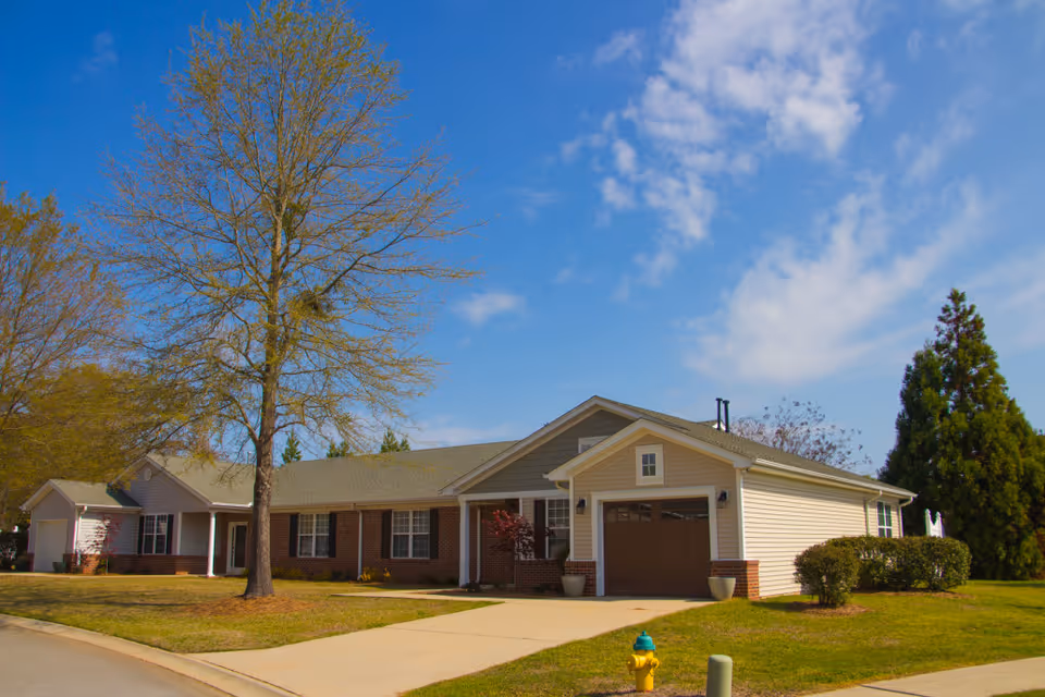 Exterior view of a single-story residential building with a combination of brick and siding walls, a garage, and a driveway. The building is surrounded by a lawn with a large tree in front and a clear blue sky with some clouds overhead.