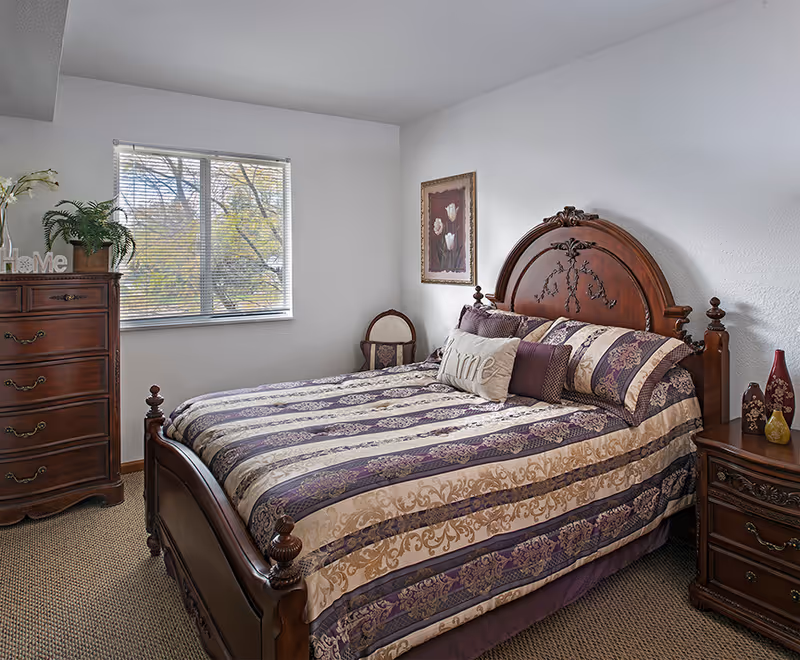 A furnished bedroom with an ornate wooden bed and patterned bedding, matching nightstands and dresser, a window, and wall art.