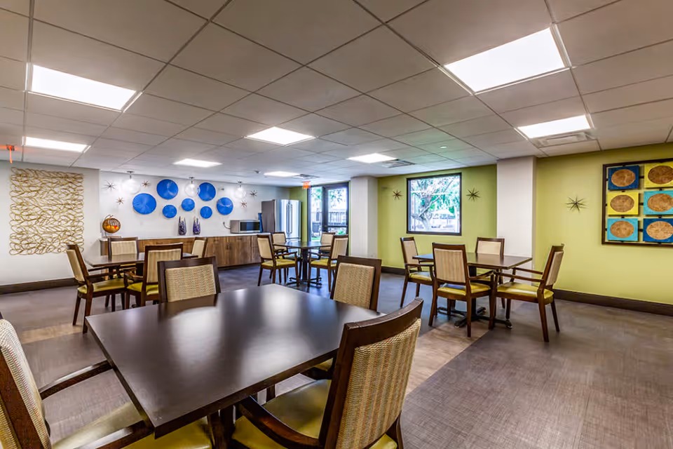 A bright dining room with several wooden tables and chairs with green cushions. The walls are decorated with blue and gold circular art pieces and starburst wall decorations. There is a kitchenette area with a refrigerator, microwave, and cabinets along one wall. Large windows let in natural light, and the ceiling has recessed lighting panels.