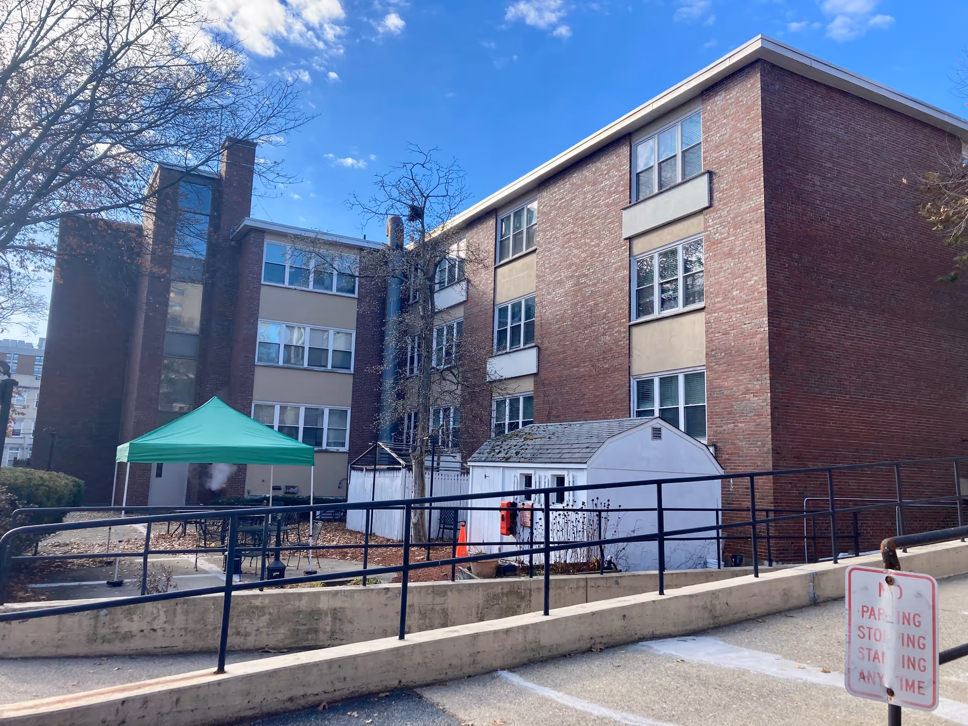 Exterior view of a multi-story brick building with several windows. In front of the building, there is a small white shed and a green canopy tent set up over a patio area with tables and chairs. A black metal railing runs along a concrete ramp leading up to the building. Leafless trees and a clear blue sky are visible in the background.