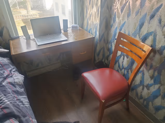 Small bedroom corner with a wooden desk by a window holding a laptop and speakers, next to a wooden chair with a red seat and a bed edge.