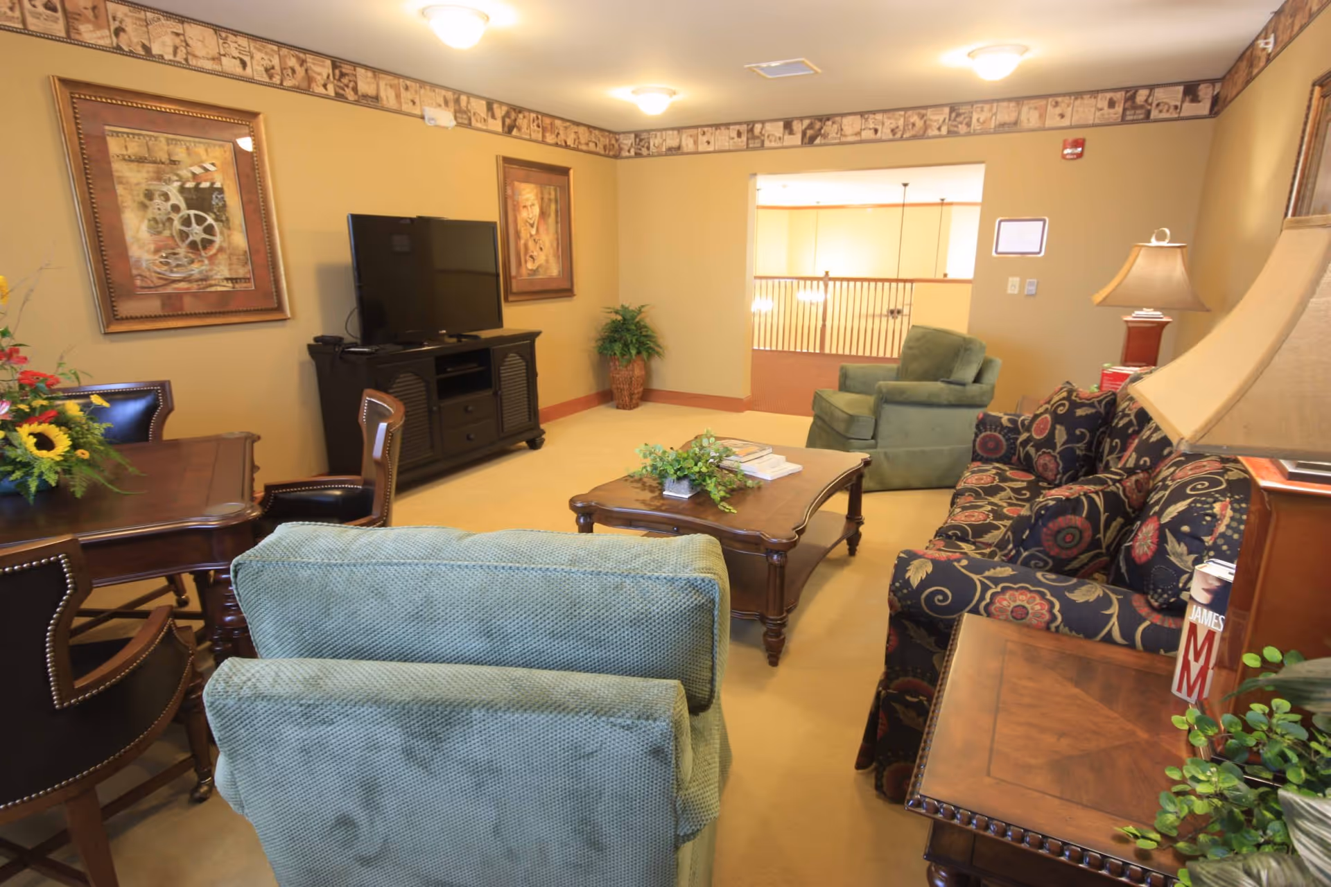A cozy living room with a patterned sofa, two green armchairs, a wooden coffee table with a plant and books, a wooden side table with a lamp and books, a TV on a black TV stand, framed artwork on the walls, and a wooden dining table with chairs. The room has beige walls with a decorative border near the ceiling and carpeted floor.