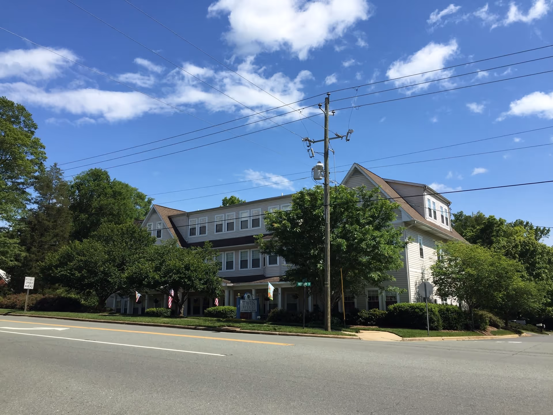 Exterior view of a three-story senior living facility building named Twin Rivers Independent Senior Living, surrounded by trees and greenery under a blue sky with some clouds. The building is located at a street corner with visible power lines and a road sign.