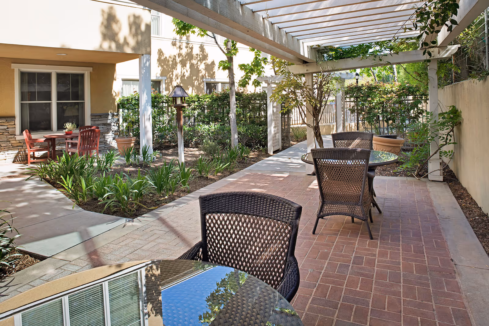 Outdoor patio area with glass-top tables and wicker chairs under a white pergola. There are plants and shrubs along the walkway, and the building exterior with windows is visible in the background.