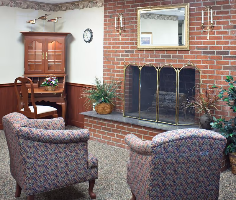 A cozy sitting area with two patterned armchairs facing a brick fireplace with a brass screen. Above the fireplace is a large mirror flanked by two wall-mounted candle holders. To the left, there is a wooden desk with a chair and a cabinet on top, decorated with a basket of flowers and three wooden bird figurines on the wall above. The room has carpeted flooring and plants placed near the fireplace.