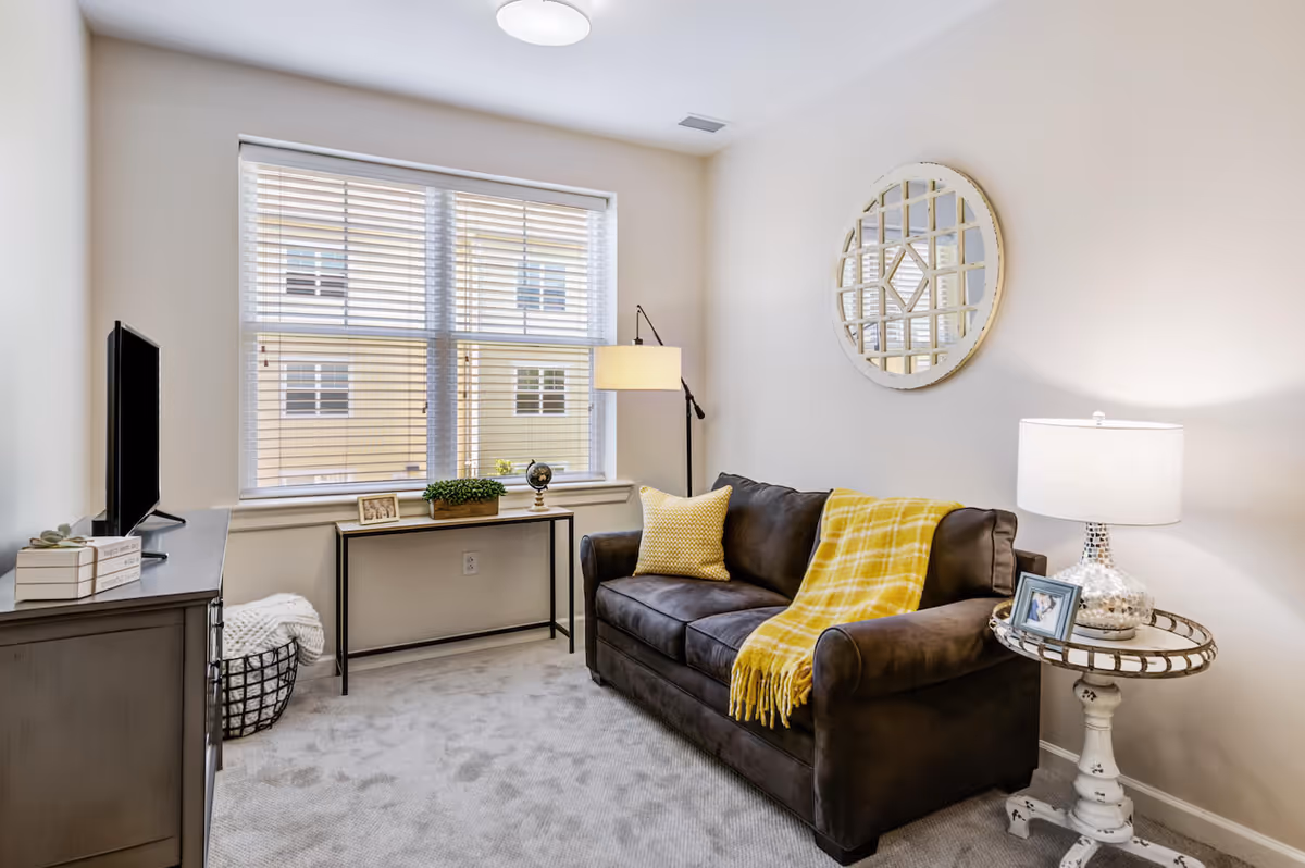 A cozy living room with a dark brown sofa adorned with a yellow pillow and a yellow plaid throw blanket. A round decorative mirror hangs on the wall above the sofa. Next to the sofa is a small white side table with a silver lamp and a framed photo. Across from the sofa is a TV on a dark wooden cabinet. A narrow console table under a large window holds a small plant, a globe, and a framed photo. The room has light-colored walls and carpeted flooring.