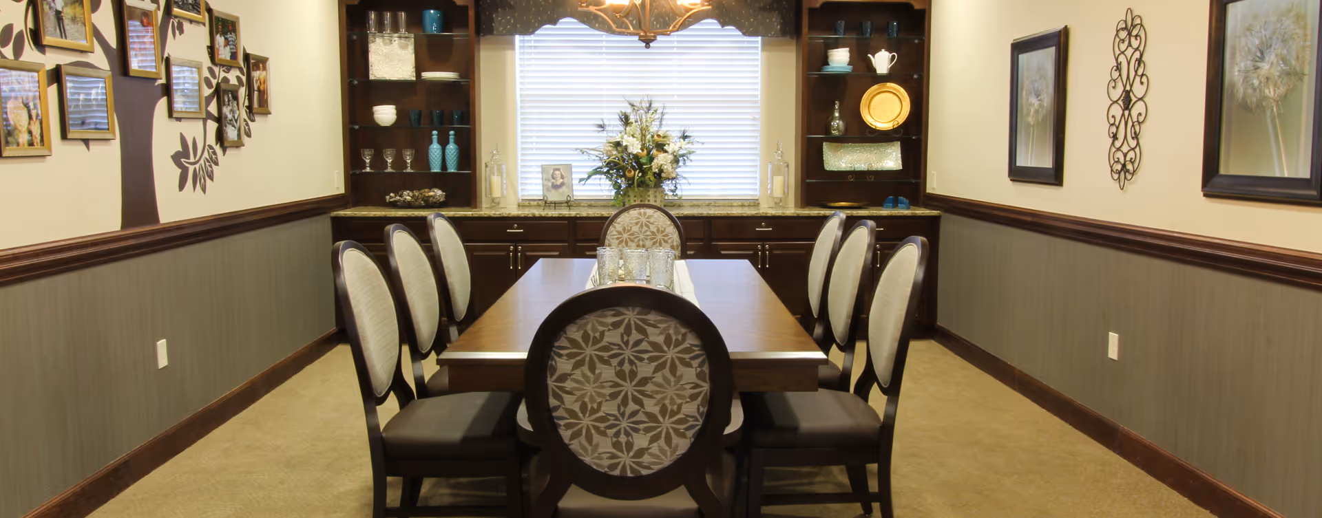 A formal dining room with a rectangular wooden table surrounded by eight upholstered chairs. The room features built-in dark wood shelves and cabinets on the far wall, decorated with glassware, plates, and ornamental items. A window with blinds and a valance is centered between the shelves, with a floral arrangement on the countertop below it. The walls are adorned with framed pictures and decorative wall art.