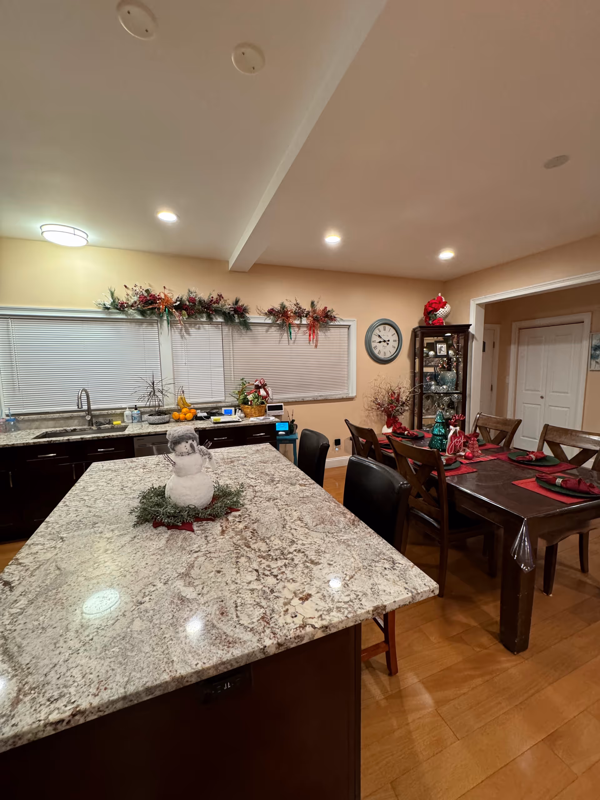 Interior view of a kitchen and dining area decorated for the holidays. The kitchen features a large granite island with a small snowman decoration on top. Behind the island are dark wood cabinets, a sink, and a window with closed blinds and festive garlands above. The dining area has a wooden table set with red placemats, napkins, and holiday decorations. A clock and a glass display cabinet with decorative items are visible on the wall.