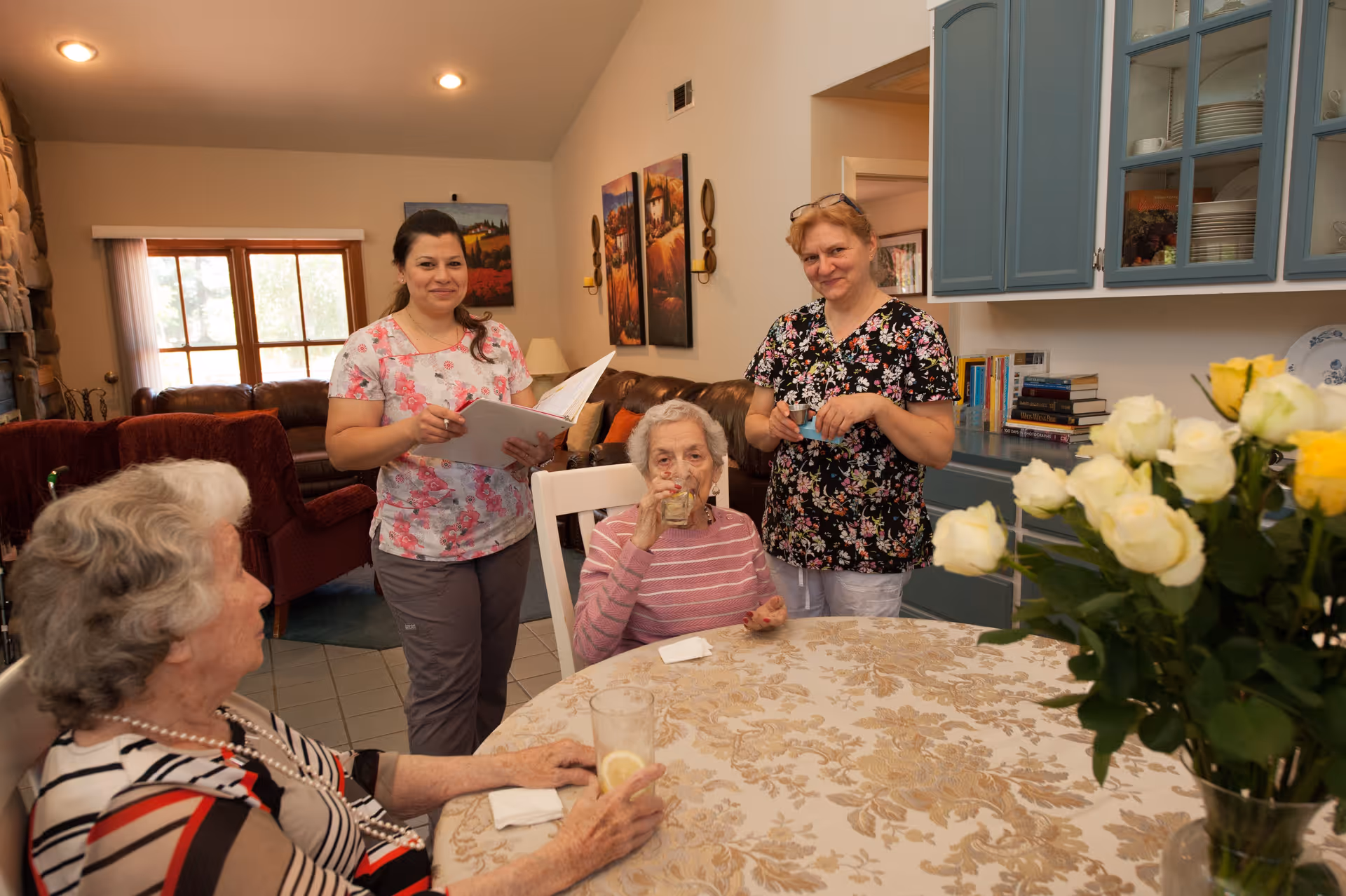 Two elderly women sitting at a round table with a floral tablecloth, one drinking from a glass and the other holding a glass with lemon. Two caregivers stand behind them, one holding a notebook and the other holding a cup. The room has a cozy living area with a window, paintings on the wall, and blue kitchen cabinets with dishes and books on the counter. A bouquet of white and yellow roses is visible on the table.