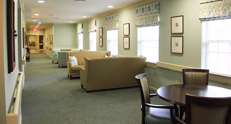 A long hallway with multiple seating areas featuring beige and light blue sofas and armchairs. There is a round wooden table with chairs near the windows on the right side. The walls are painted light green and decorated with framed pictures and patterned window valances. The floor is carpeted in a muted green tone.