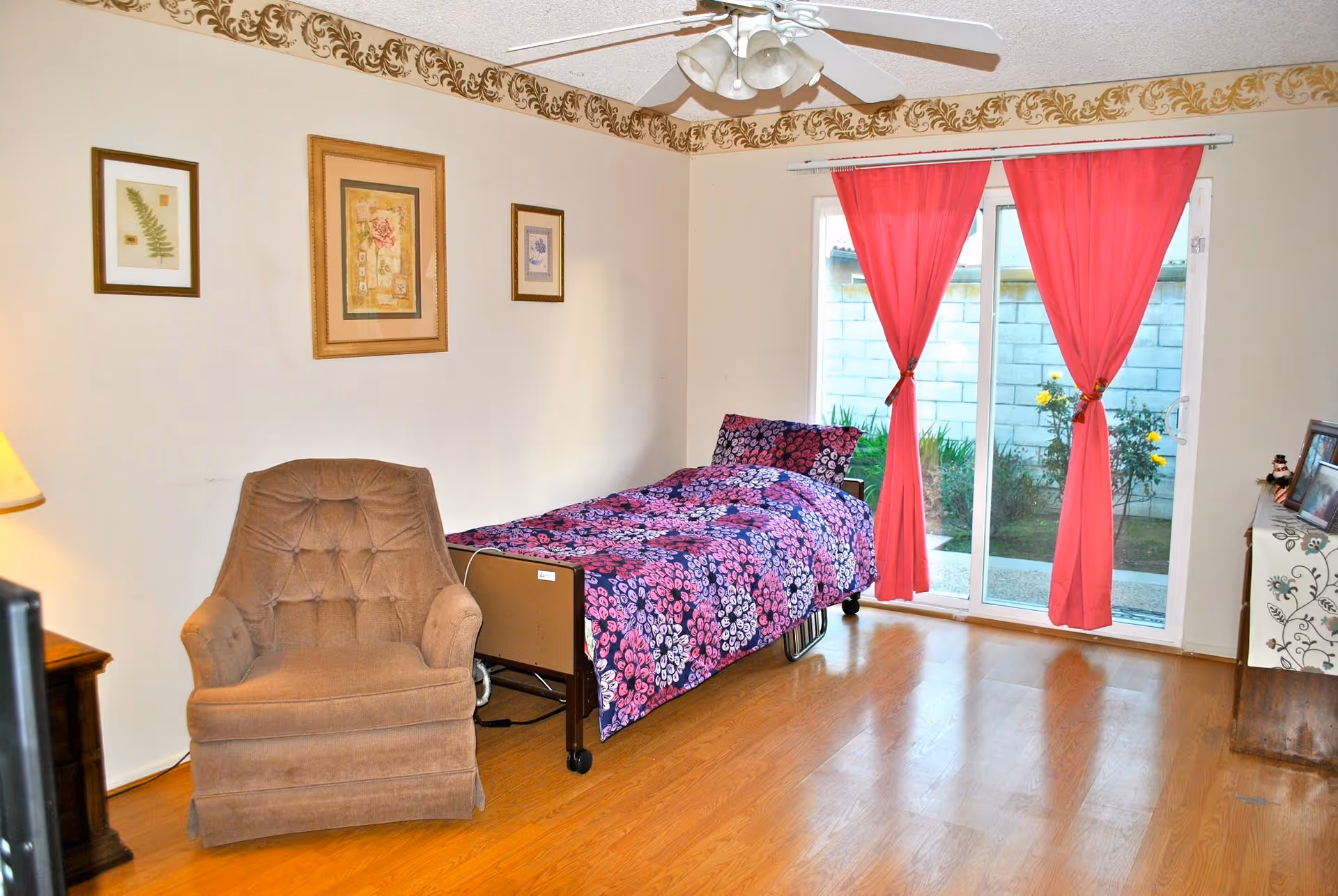 A bedroom with a single bed covered in a purple and pink floral bedspread, a brown upholstered armchair, three framed pictures on the wall, a ceiling fan with lights, and sliding glass doors with red curtains leading to an outdoor garden area.
