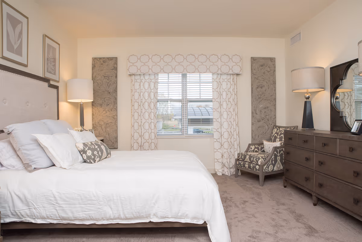 Well-lit bedroom with a made bed, patterned armchair, large dresser and a window with patterned curtains.