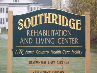 A green and gold sign for Southridge Rehabilitation and Living Center, a North Country Health Care Facility, with additional signs below for Residential Care Services and Outpatient Therapy. The sign is outdoors with a grassy area and houses in the background.