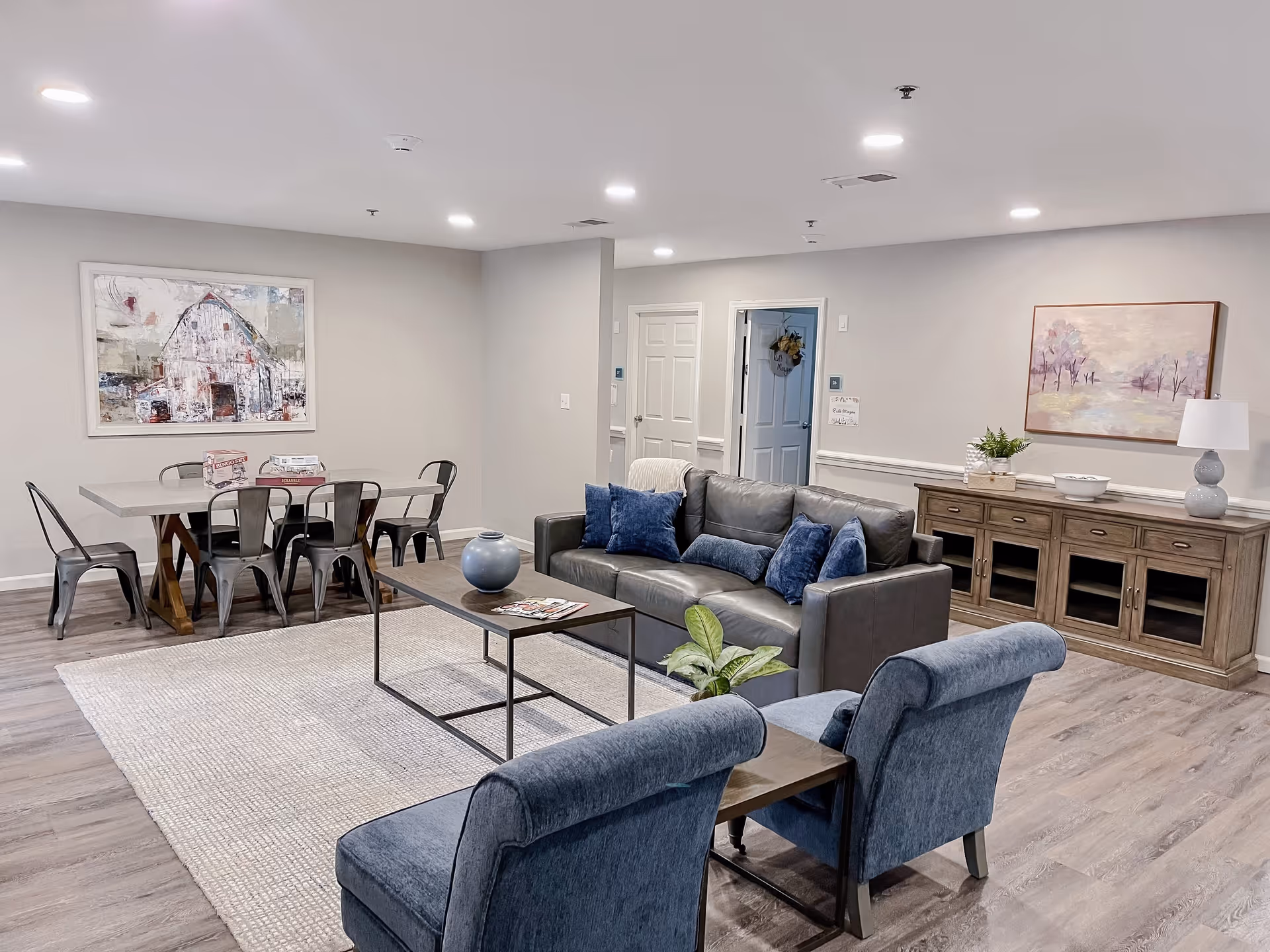 A modern living room and dining area in a senior living facility. The living room features a gray leather sofa with blue cushions, two blue upholstered chairs, a wooden coffee table with a decorative vase, and a light-colored rug. The dining area has a rectangular table with six metal chairs. The walls are light gray with two framed paintings, one depicting a barn and the other a landscape. A wooden sideboard with a lamp, plant, and bowl is against the wall. The room has recessed ceiling lights and wood-style flooring.