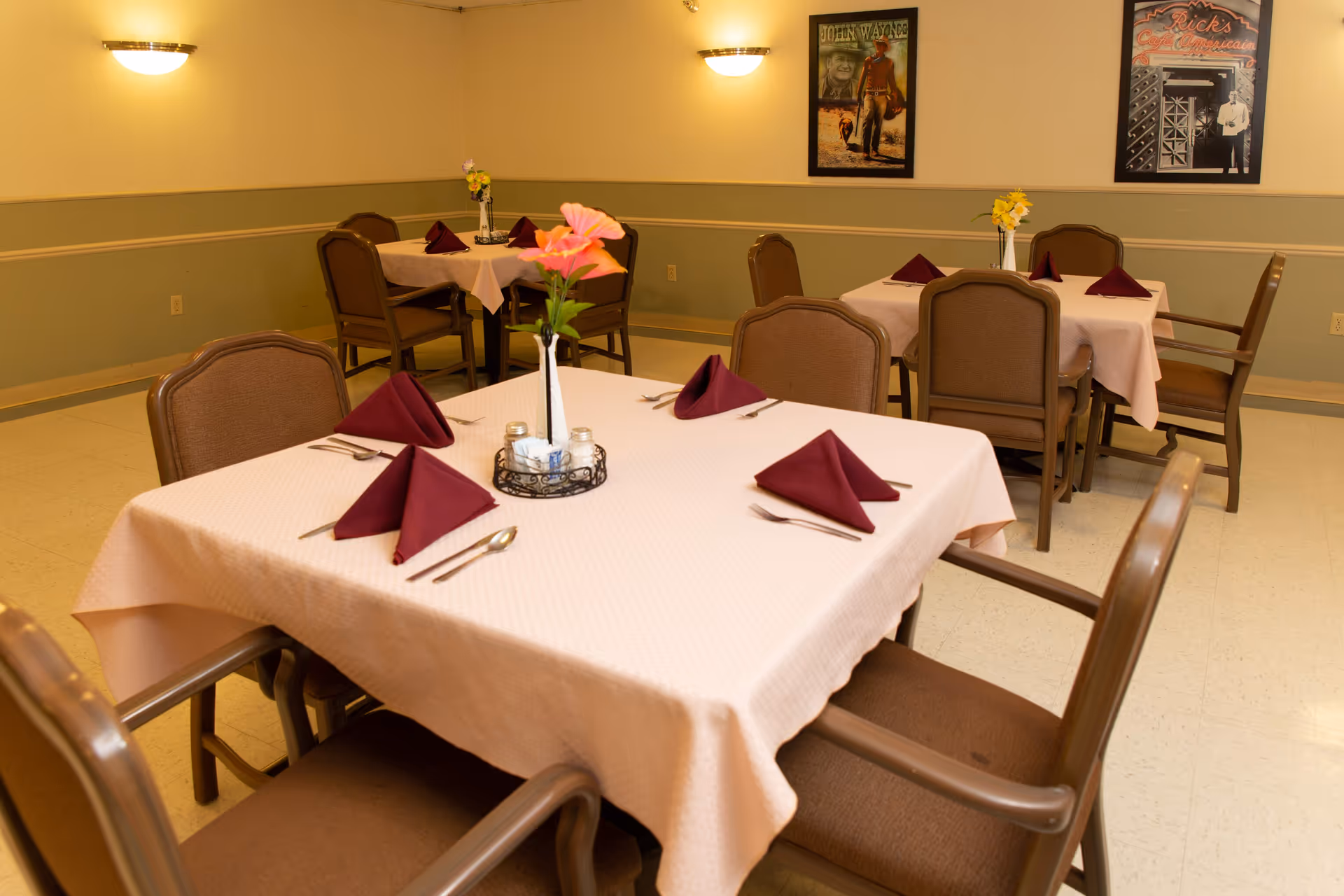 Dining room with tables covered in light pink tablecloths, each set with maroon folded napkins, silverware, and a small vase with flowers. Brown cushioned chairs surround the tables. The walls are painted beige and green with two framed vintage posters hanging on the wall and wall-mounted lights providing illumination.