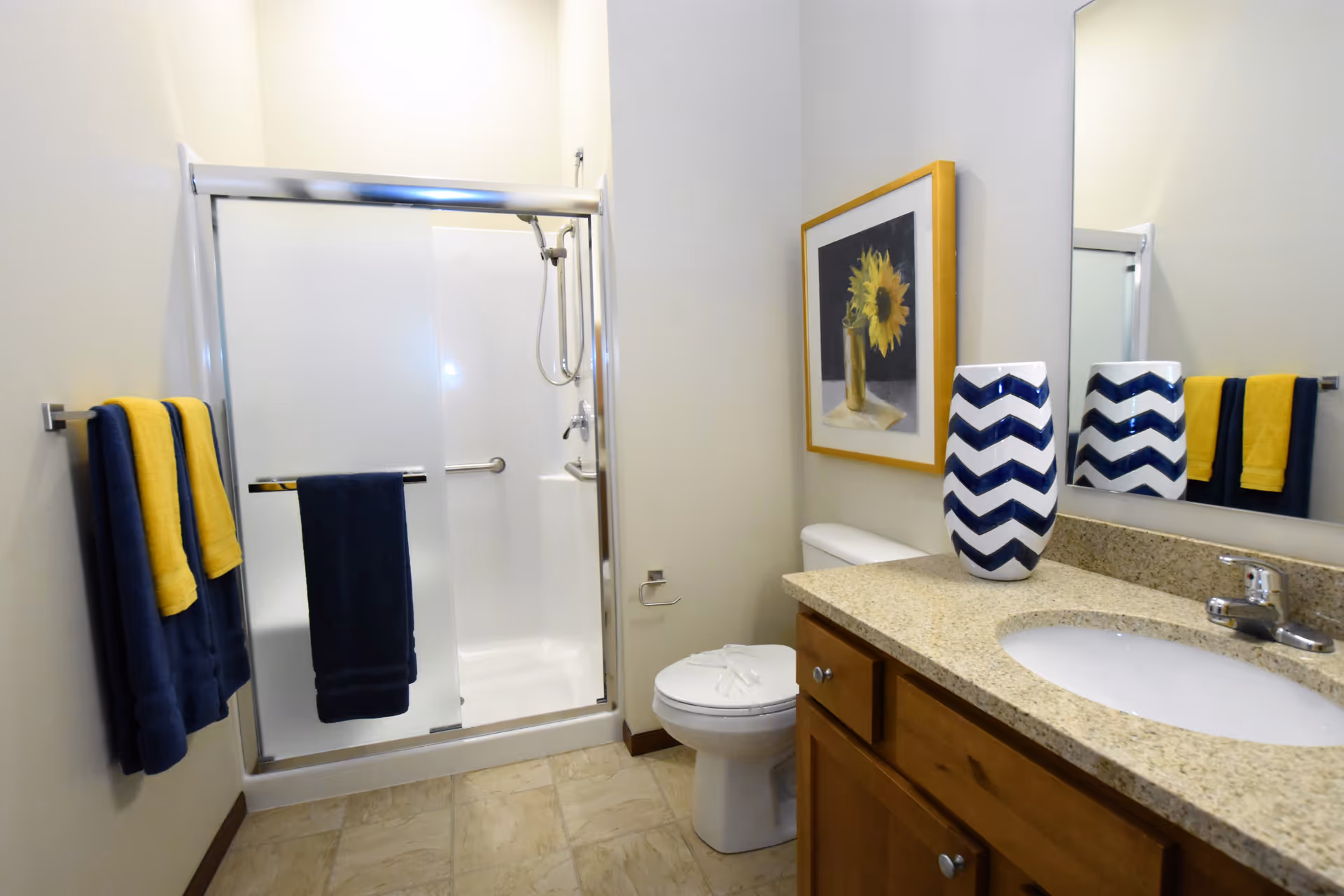 Bathroom with a glass-enclosed shower, toilet, vanity with sink and mirror, and blue and yellow towels.