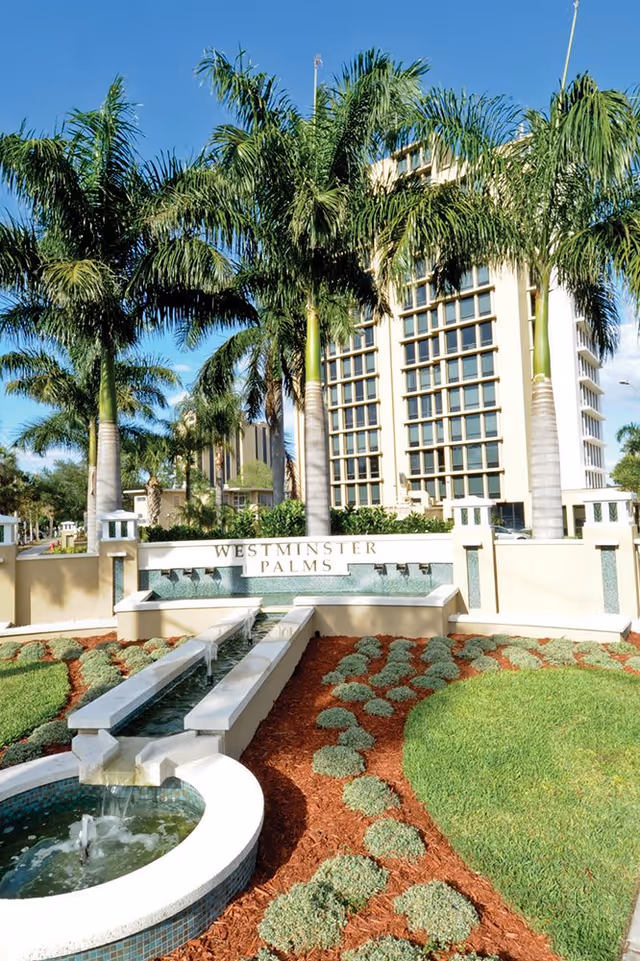 Outdoor view of Westminster Palms facility entrance with a water feature, landscaped garden with small bushes and mulch, tall palm trees, and a multi-story building in the background under a clear blue sky.