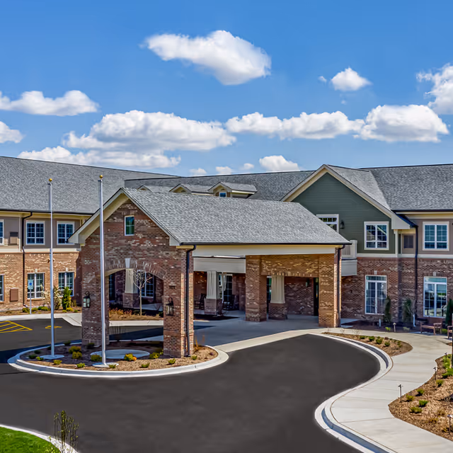 Exterior view of American House Mount Prospect, showing a large brick and siding building with a covered entrance driveway, multiple windows, and a clear blue sky with scattered clouds above.