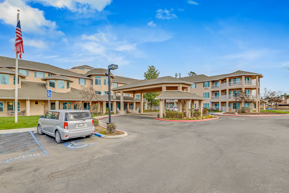 Exterior view of Solstice Senior Living at Bakersfield showing a multi-story building with beige walls and multiple windows. There is a covered entrance with a driveway and a silver car parked in a handicapped parking space. An American flag is flying on a flagpole to the left, and the sky is partly cloudy.