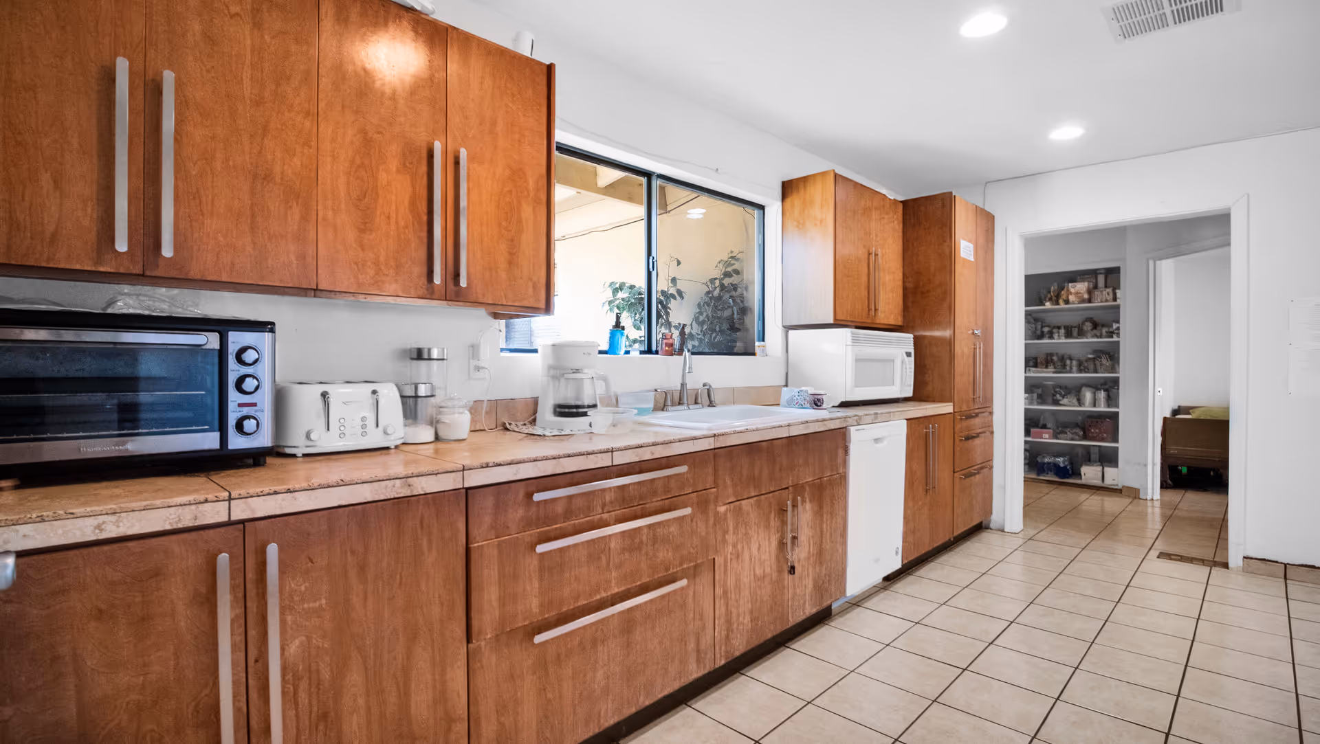 A kitchen with wooden cabinets and drawers, a tiled floor, and a window above the sink. On the countertop, there is a toaster oven, a toaster, a coffee maker, and a microwave. The kitchen opens into a hallway with shelves stocked with various items.