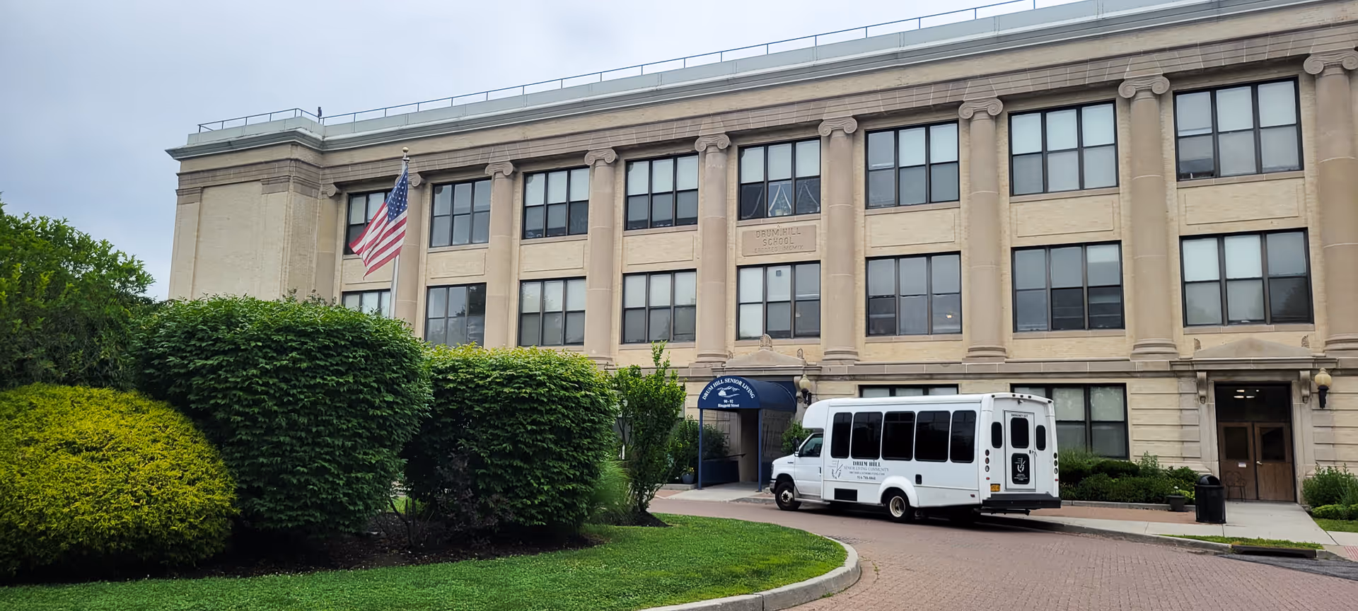 Front exterior of a three-story beige senior living building with an entrance canopy, landscaped shrubs, an American flag, and a parked white shuttle van.