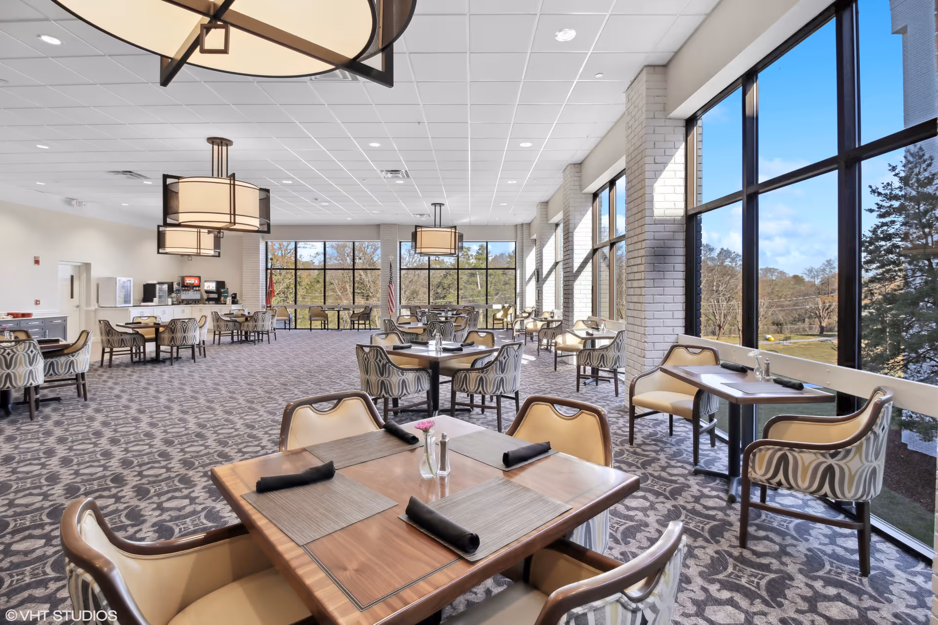 A spacious dining room with large windows allowing natural light to fill the room. The room features multiple wooden tables with beige and patterned chairs, each table set with placemats and black napkins. Modern pendant lights hang from the ceiling, and there is a beverage station along one wall. Outside the windows, trees and a clear blue sky are visible.