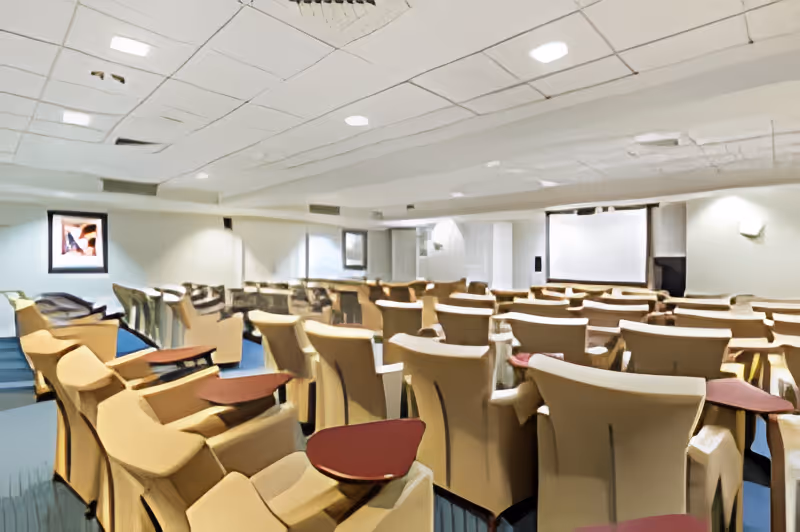A theater-style meeting room with rows of beige reclining chairs each with small attached red tray tables facing a projection screen.