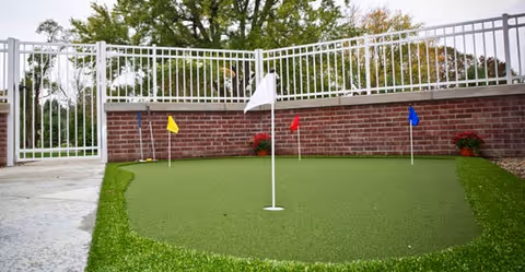 A small outdoor putting green with four colored flags (white, yellow, red, blue) surrounded by a white metal fence and a brick wall, with trees visible in the background.