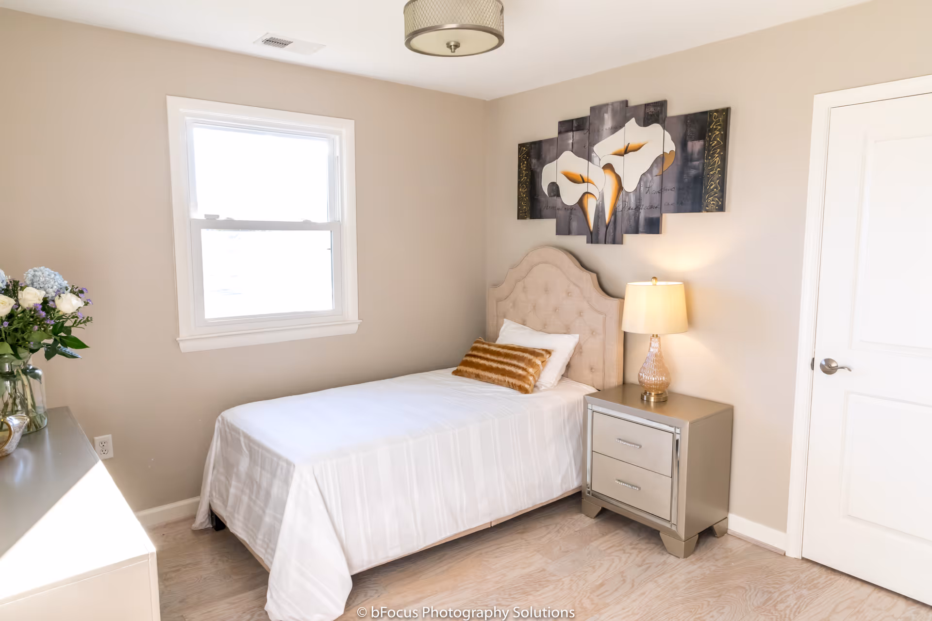 A bright and clean bedroom with a single bed covered in white linens, a beige tufted headboard, and a decorative brown pillow. Next to the bed is a gray nightstand with two drawers and a lamp with a beige shade. Above the bed hangs a multi-panel artwork featuring white flowers. There is a window on the left wall letting in natural light, and a white door on the right side of the room. A vase with flowers is partially visible on a dresser to the left.