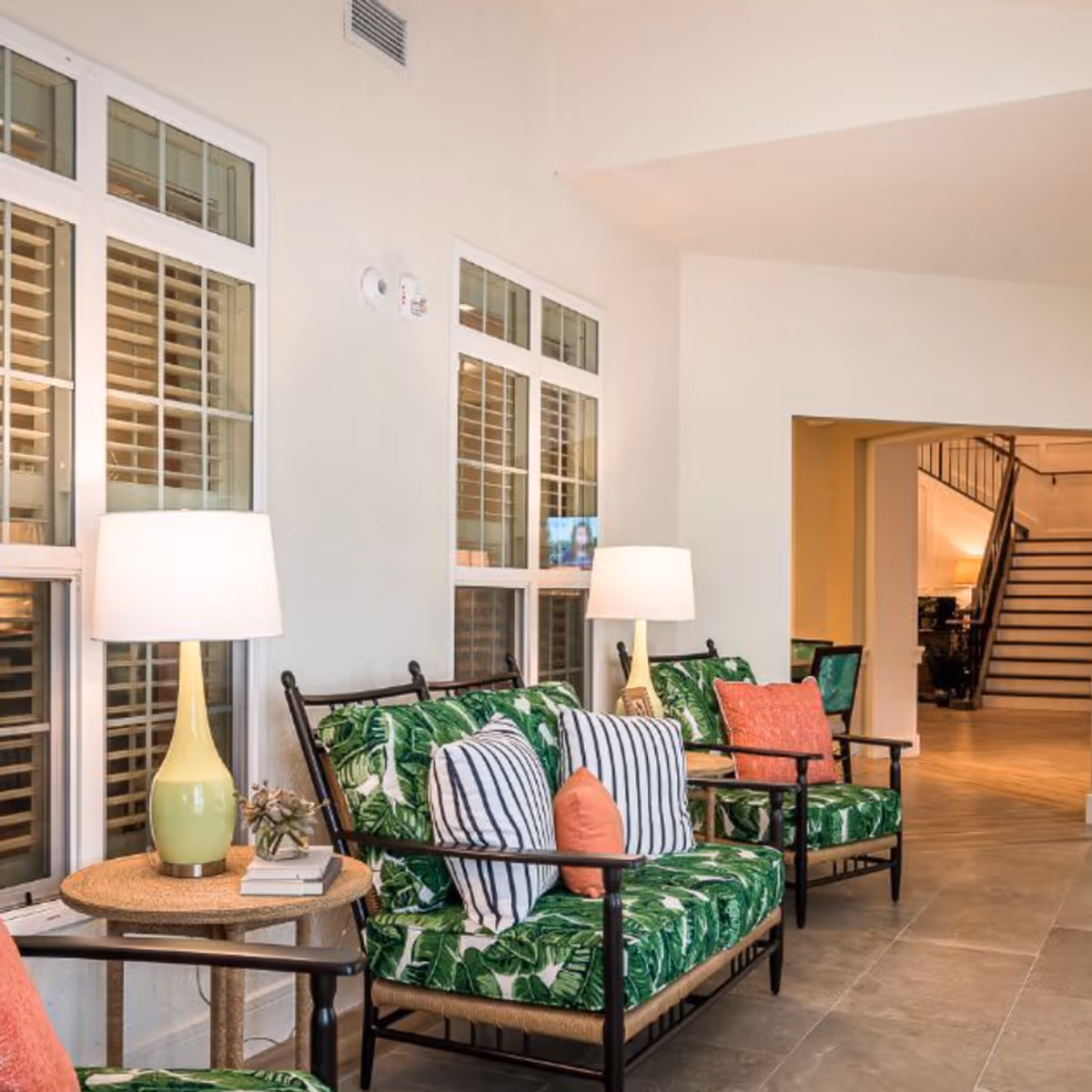 A bright and inviting interior seating area with two chairs and a loveseat featuring green leafy patterned cushions and colorful throw pillows. Two yellow table lamps sit on round wicker side tables next to the seating. Large windows with white frames and shutters are visible behind the furniture. In the background, there is a hallway leading to a staircase with black railings.