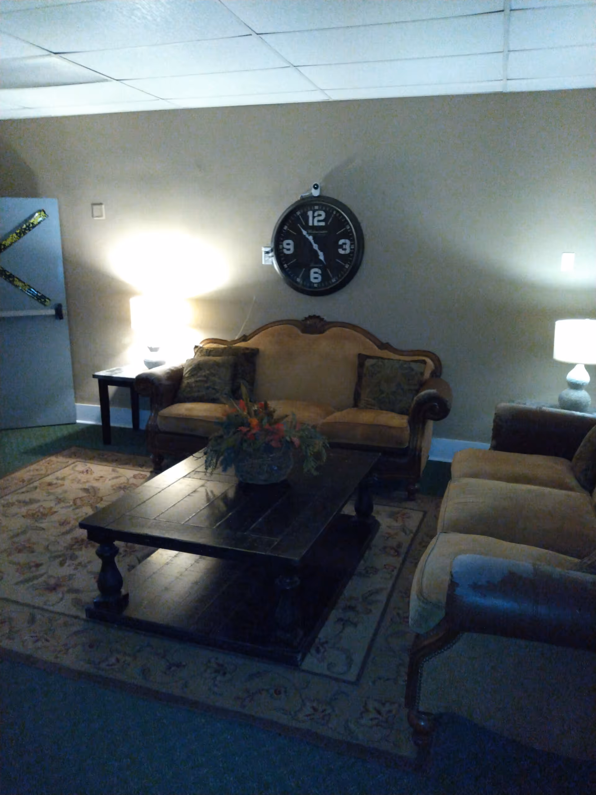 A cozy living room with two beige upholstered sofas, a dark wooden coffee table with a floral arrangement, two side tables each with a lamp, a large wall clock above one sofa, and a patterned area rug on the floor.