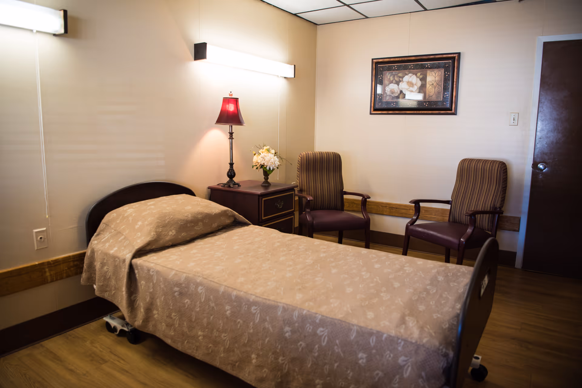 A simple senior bedroom with a single bed, nightstand topped with a lamp and flowers, and two striped chairs against the wall.