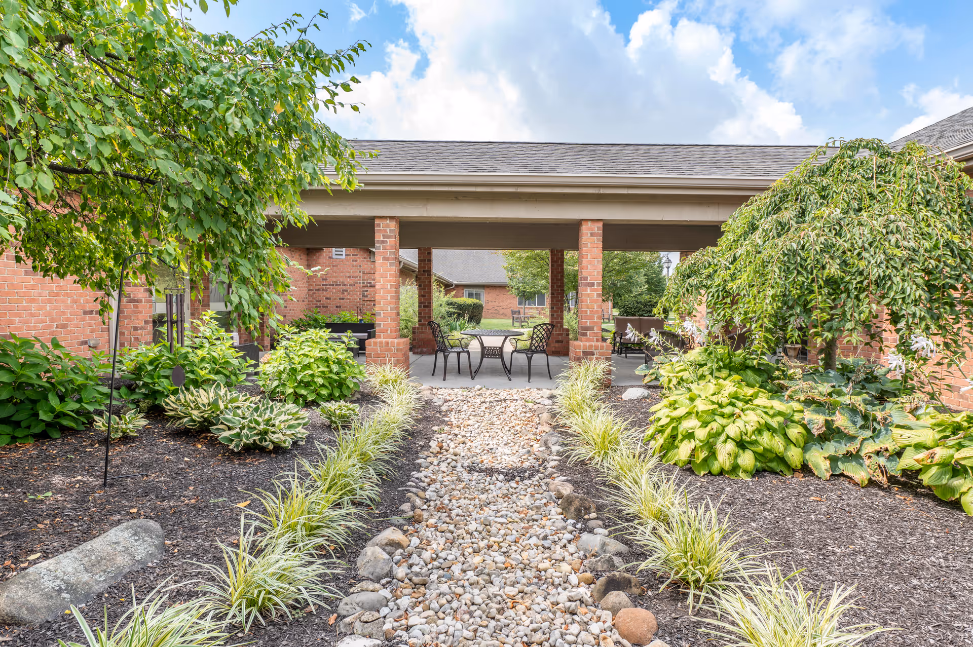 Outdoor garden area with a stone pathway lined by plants and shrubs leading to a covered patio with chairs and tables. The area is surrounded by brick buildings and greenery under a partly cloudy sky.