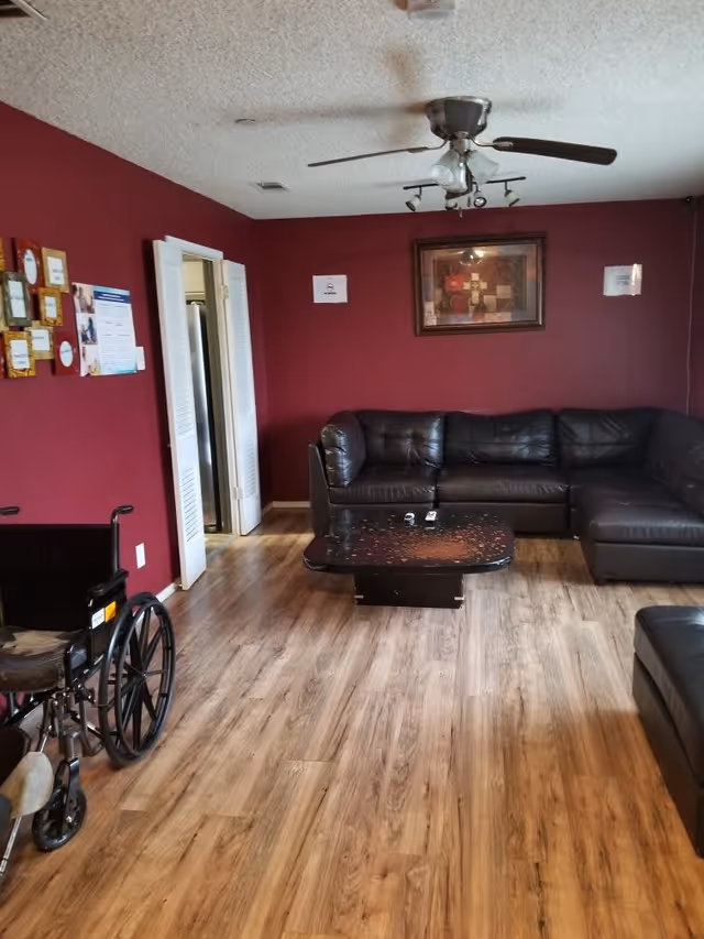 A living room with dark red walls and wood flooring. There is a black leather sectional sofa along the right wall, a black coffee table in front of it, and a ceiling fan with lights above. A wheelchair is positioned on the left side near the entrance to another room with white double doors. Various framed pictures and notices are hung on the left wall.