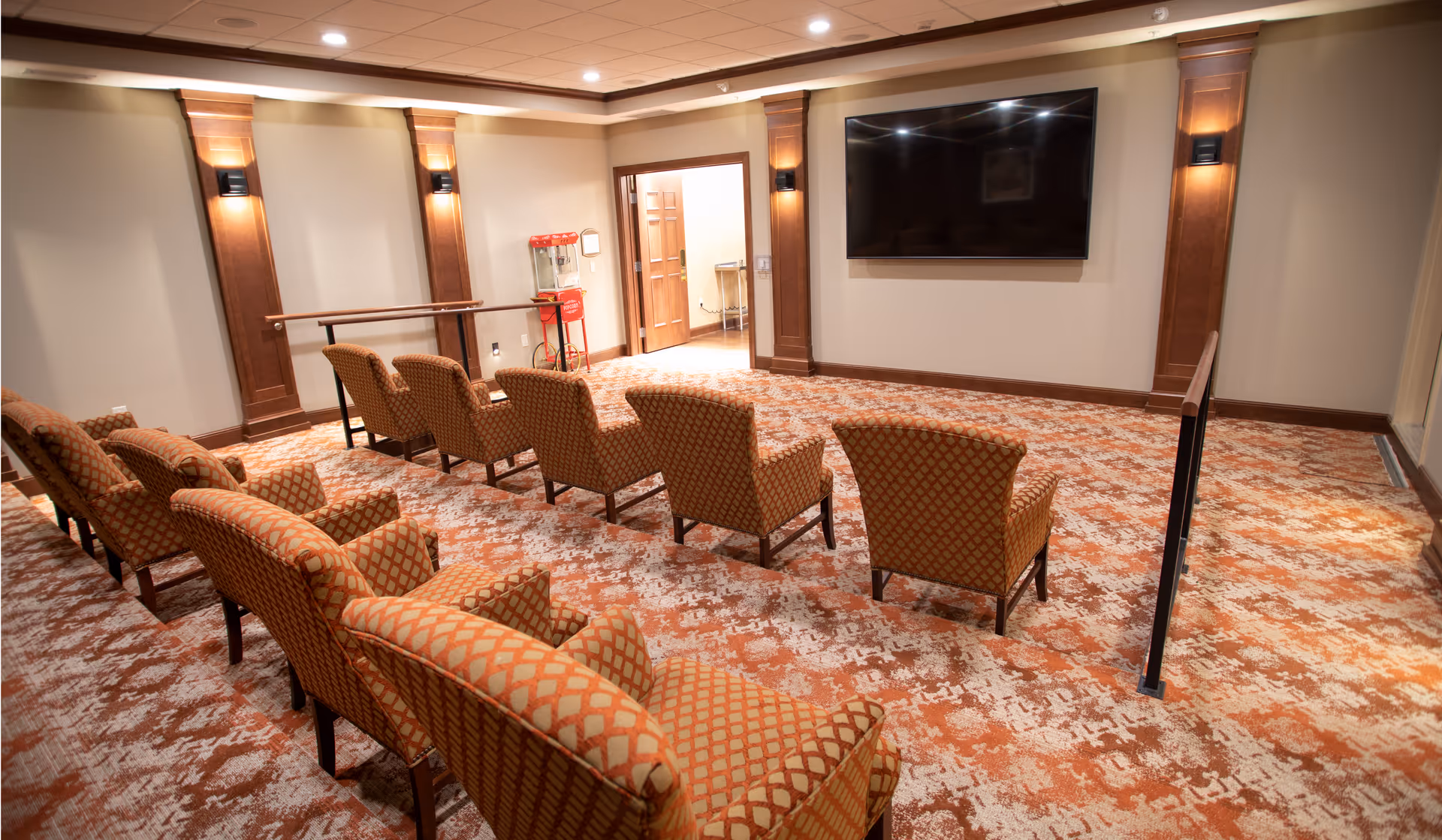 A small theater room with two rows of patterned upholstered chairs facing a large flat-screen TV mounted on the wall. The room has warm lighting with wall sconces, wood panel accents, and a patterned carpet in shades of orange and beige. There is a popcorn machine near the doorway at the back of the room.