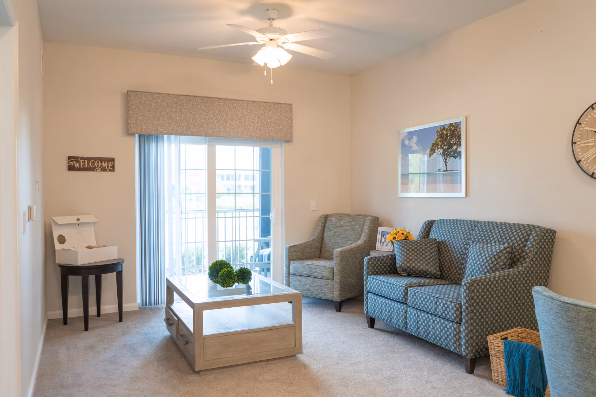 A cozy living room with a patterned loveseat and armchair, a wooden coffee table with decorative greenery, a small side table with a flower arrangement and framed photo, a large wall clock, and a framed picture on the wall. Sliding glass doors with vertical blinds lead to an outdoor balcony.