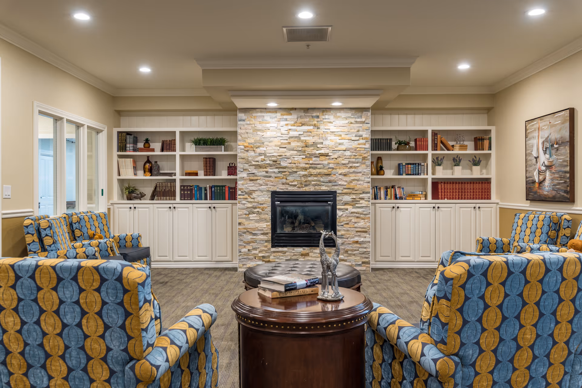 A cozy living room area with four patterned armchairs arranged around a round wooden table and a circular ottoman. The room features a stone fireplace centered between two white built-in bookshelves filled with books and decorative items. The walls are painted beige, and there is a painting of sailboats on the right wall. The ceiling has recessed lighting.