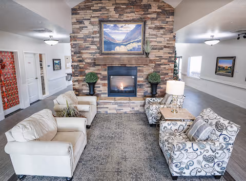 Communal living room with upholstered chairs arranged around a stone fireplace topped by a landscape painting.