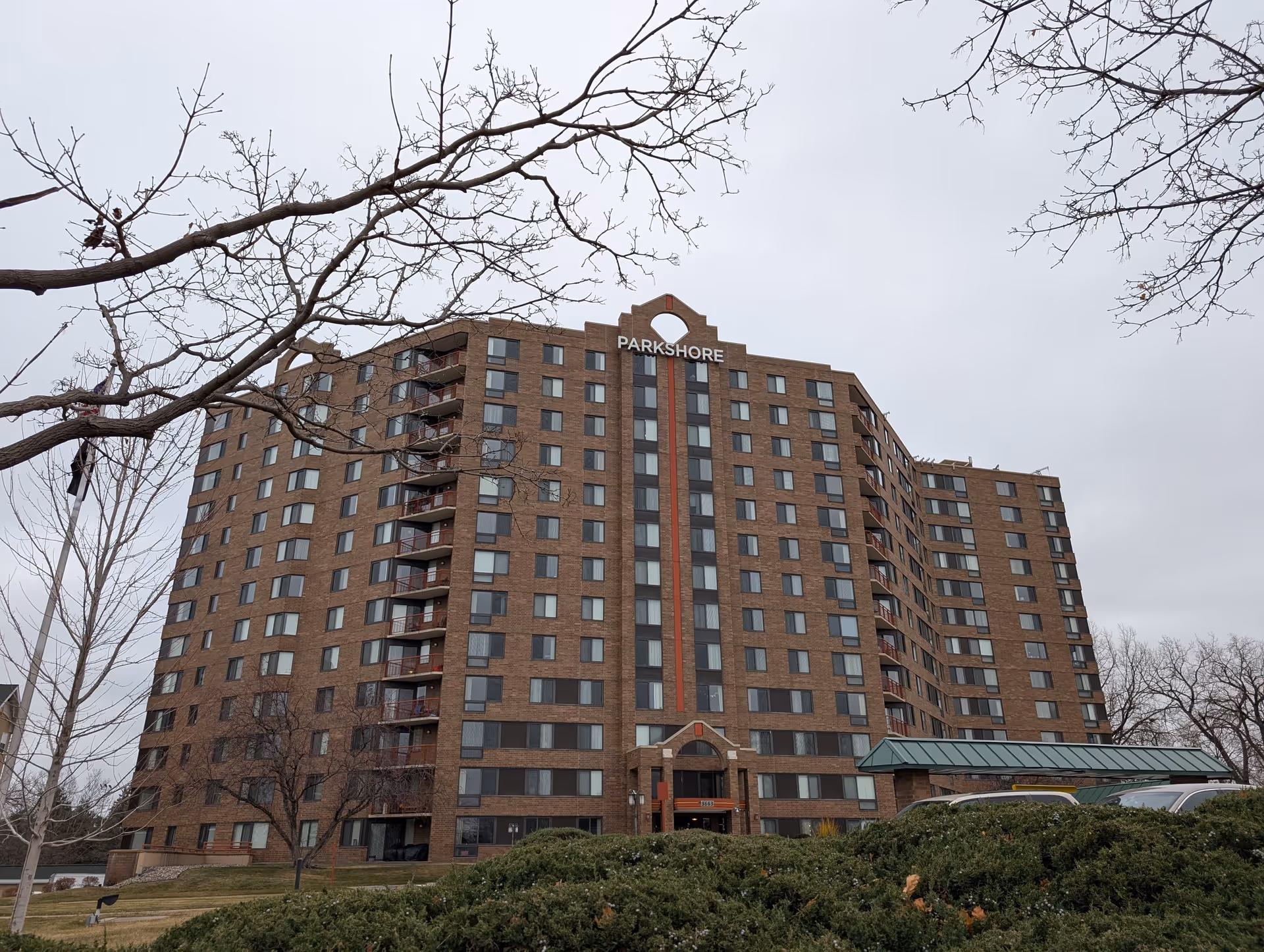 Large brick multi-story senior living building with a 'Parkshore' sign above the main entrance, framed by bare trees.