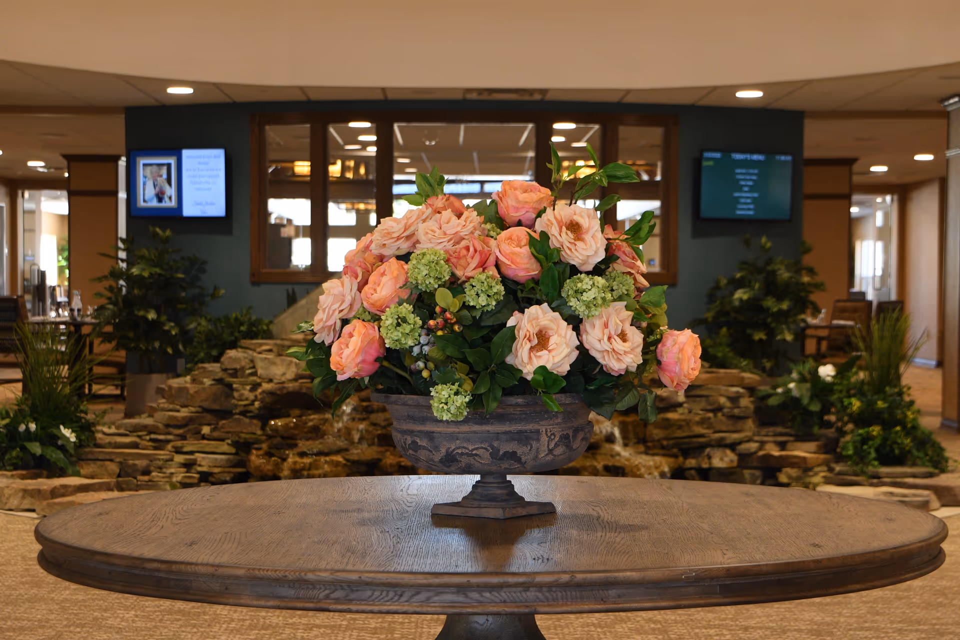 A large floral arrangement with pink and peach roses and green hydrangeas in a decorative vase sits on a round wooden table in the center of a spacious indoor area. Behind the table, there is a rock water feature with plants and two digital screens mounted on the walls. The setting appears to be a welcoming common area inside a senior living facility.