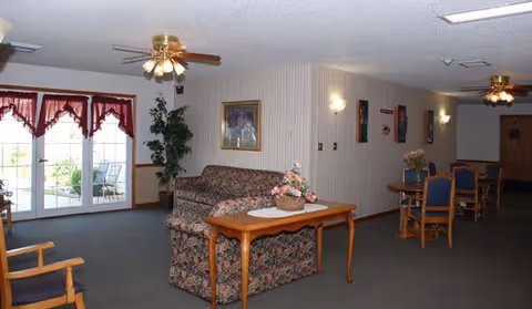 Interior view of a senior living facility common area with floral patterned sofa, wooden coffee table with a flower basket, wooden chairs, and tables. The room has ceiling fans with lights, framed pictures on the walls, and large glass doors with red curtains leading outside.