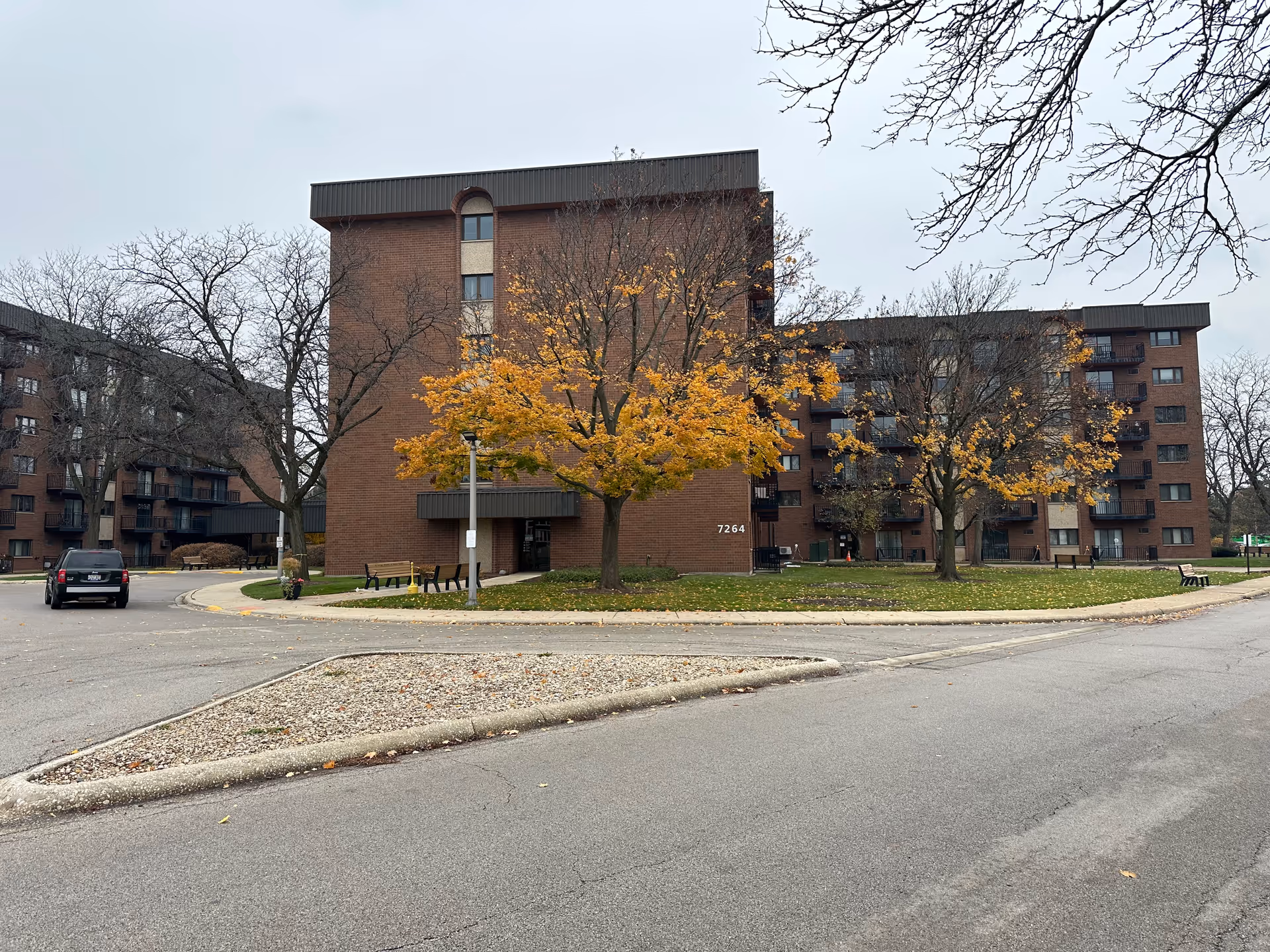 Exterior view of a multi-story brick building with balconies, surrounded by trees with autumn foliage and a paved driveway. A black SUV is parked on the left side near the building entrance, and there are benches and a street lamp near the entrance. The building number 7264 is visible on the wall.
