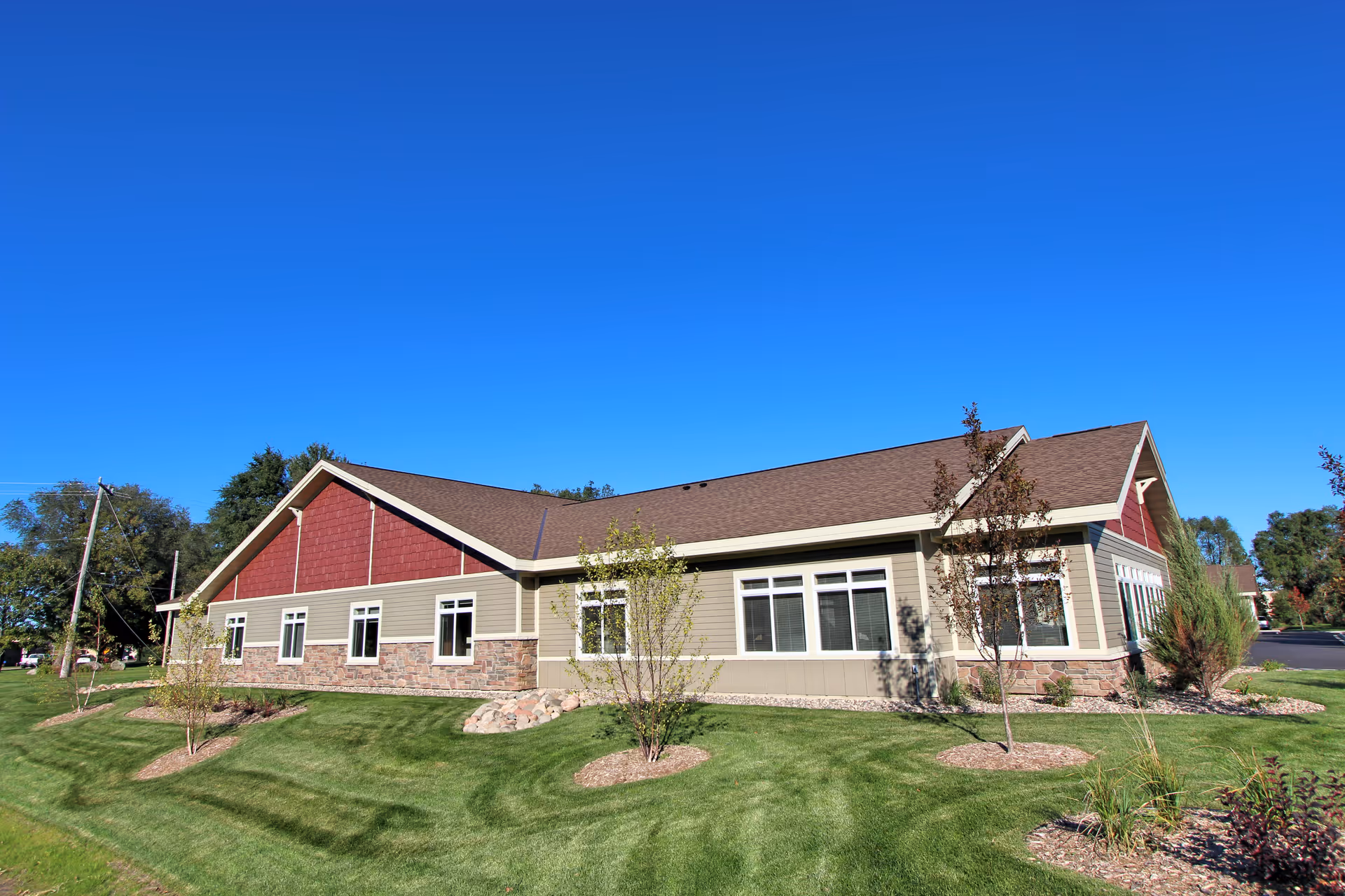 Exterior view of a single-story building with beige siding, red gable accents, and multiple windows under a clear blue sky. The building is surrounded by neatly maintained green grass and small trees.