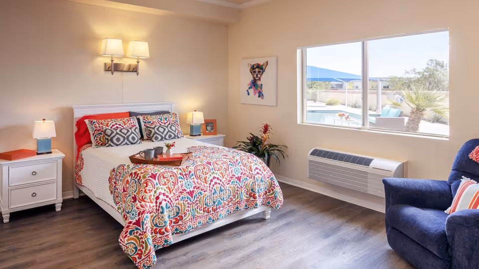 A bright bedroom with a white bed frame and colorful patterned bedding. There are two white nightstands with lamps on either side of the bed. A tray with cups and a small flower vase is placed on the bed. A blue armchair with a striped pillow is positioned near a large window that shows an outdoor pool area with palm trees. The room has wooden flooring and a wall-mounted air conditioning unit below the window. A colorful dog artwork hangs on the wall next to the window.