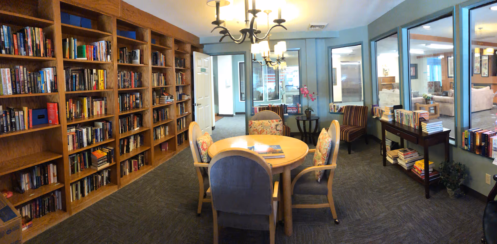 Cozy interior library/common room with built-in bookshelves along the left wall, a round table and chairs in the center, and additional seating by windows overlooking an adjacent lounge.