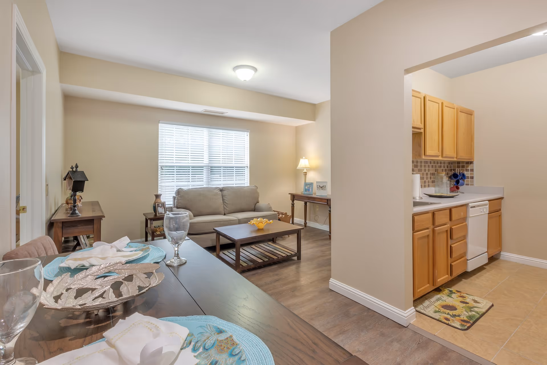 Open-plan living area with a sofa and coffee table, dining table in the foreground, and a small kitchen with wooden cabinets to the right.