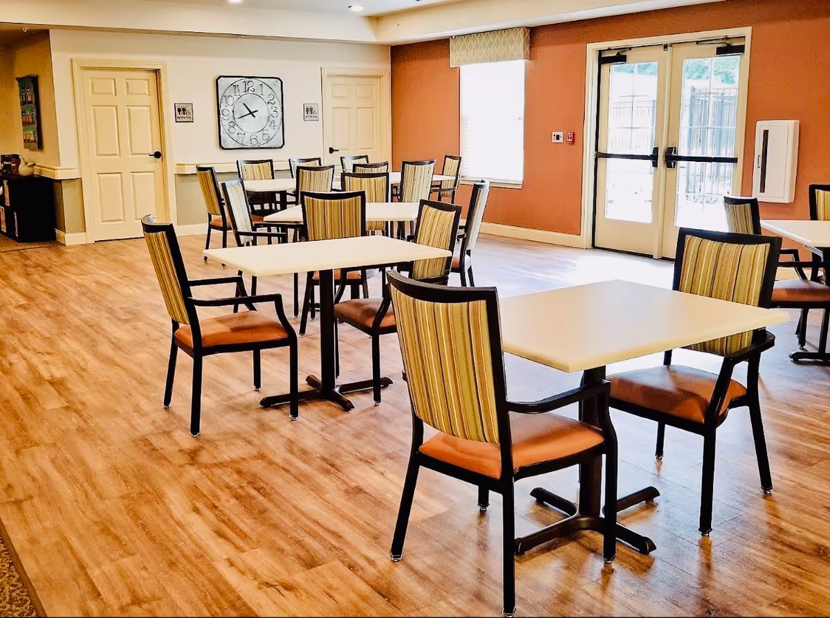 A bright dining room with several square tables and striped cushioned chairs arranged neatly on a wood floor. The room has light-colored walls with two closed doors, a large clock on the wall, a window with a valance, and double glass doors leading outside.