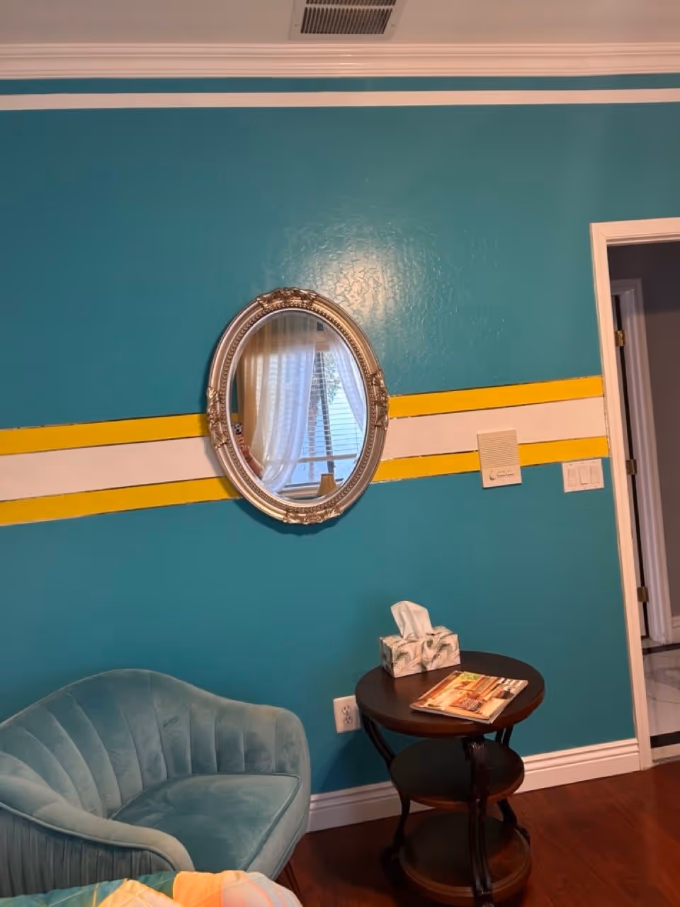 A cozy corner of a room with teal walls featuring a horizontal white and yellow stripe. An ornate oval mirror hangs on the wall reflecting a window with sheer curtains. Below the mirror is a small round wooden side table with a tissue box and a magazine on it. To the left of the table is a plush teal armchair. A doorway is visible on the right side of the image.